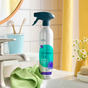 Grove Co.s Foaming Bathroom Cleaner in a recyclable aluminum spray bottle is on a bathroom counter by the sink, next to a green cloth, toothbrushes, toothpaste holder, and a small vase of daisies. A mirror and tiled wall are visible in the background.
