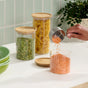 A hand pours red lentils from a metal measuring cup into a Grove Co. Bamboo & Glass Storage Jar on the kitchen counter, surrounded by jars filled with green split peas and spiral pasta, plus stacked green bowls.