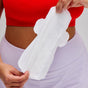 A person wearing a red top and white bottoms holds an unused Cora Made to Move Pad in front of their torso, highlighting its You-Shaped™ fit for reliable leak protection.