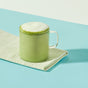 A glass mug of blume Organic Matcha Coconut Latte Powder sits frothy on a white and green striped cloth, atop a blue surface with a pale background.