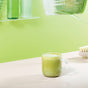 A glass mug filled with a frothy green drink made from blume Organic Matcha Coconut Latte Powder sits on a light surface, set against a pastel green background with modern decor, including bottles and a plant.