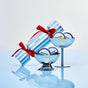 Two silver bowls hold round Everist "The Holiday Bonbon" shampoo and conditioner minis in front of an Everist blue-and-white striped gift box with a red ribbon, displayed on a light blue reflective surface. The perfect travel-friendly gift.