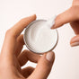 A person holds an open tin of Everist’s The Holiday Bonbon—travel mini-sized—scooping out white cream with their index finger. The background is plain and light-colored.