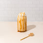 A glass of iced caramel drink made with blume Organic Salted Caramel Latte Powder is topped with whipped cream and caramel drizzle, set on a light surface with caramel drips and a gold spoon of powder nearby. White subway tile background.