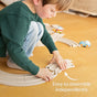 A young child in a green sweater and jeans kneels on a wooden floor, assembling the HABA Cars Expansion Set made from 100% recycled materials for ages 2+. Nearby, toy cars sit on the toddler track. Text: “Easy to assemble independently.”.