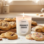 A lit Homesick Grandma's Cookies Candle sits on a kitchen counter surrounded by cookies, filling the room with a cozy, inviting aroma from this cruelty-free soy wax candle.