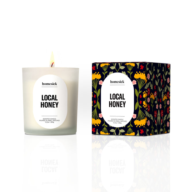A lit white candle labeled Homesick Hot Cider, made from natural soy wax, stands next to its box, which is decorated with colorful flowers and bees on a dark background.