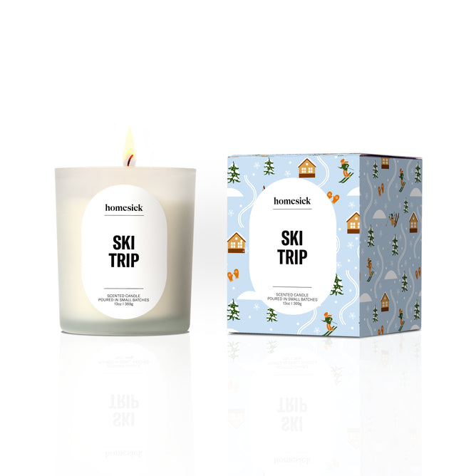 A lit Homesick Hot Cider Candle made from natural soy wax sits in a glass holder next to its decorative box, both displaying cozy autumn-inspired designs.