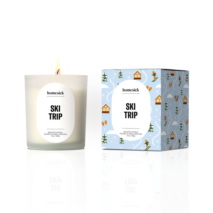 A lit Homesick Hot Cider Candle made from natural soy wax sits in a glass holder next to its decorative box, both displaying cozy autumn-inspired designs.