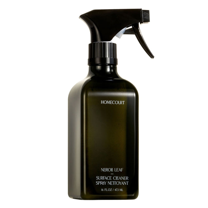 A green Homecourt Surface Cleaner spray bottle (16 fl oz/473 ml) with a black nozzle sits against a white background. The cleaner is made with skin-safe ingredients and is infused with fine fragrance.