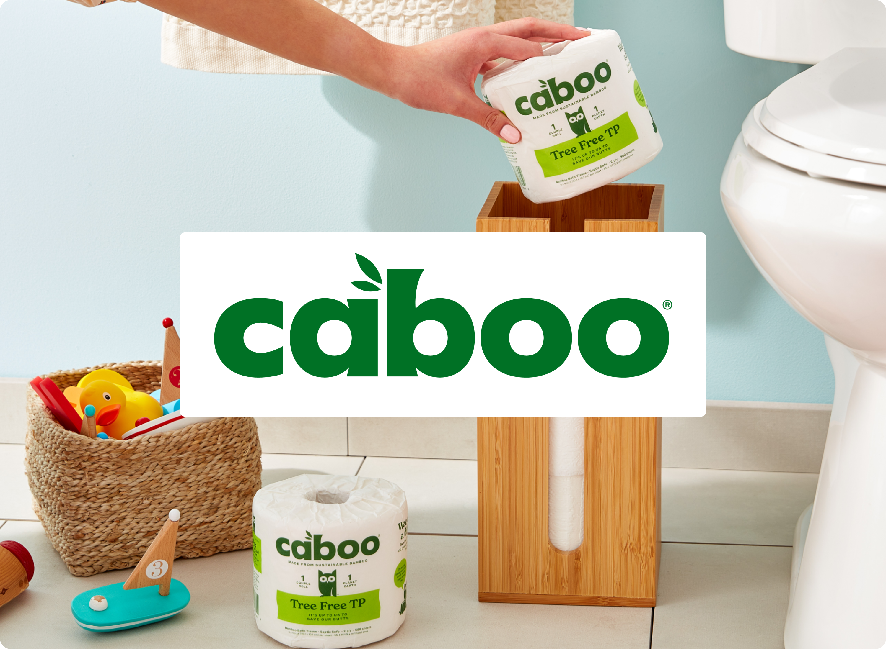 A person places a Caboo tree-free toilet paper roll into a wooden holder in a bathroom. Another roll and a basket of toys are nearby. The Caboo logo is prominently displayed in the center.