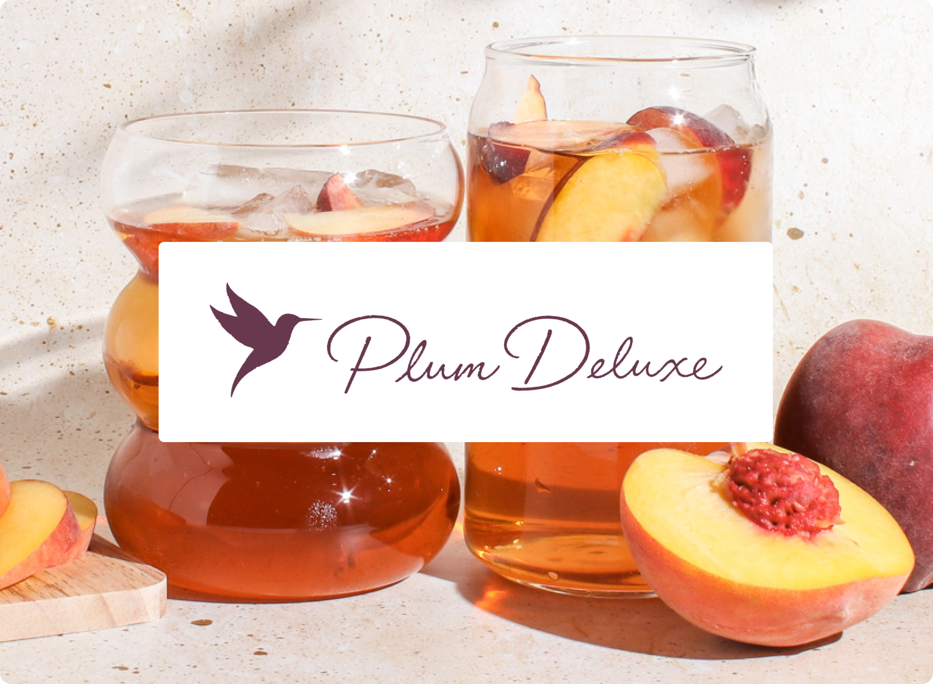 Two glasses of fruit punch with a 'Plum Deluxe' label, surrounded by peaches and raspberries on a light background.