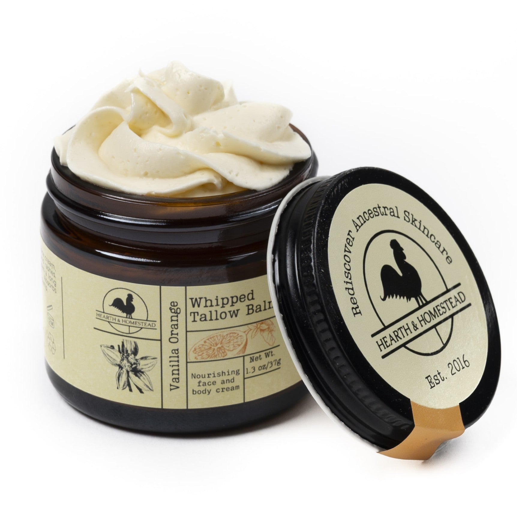 An open jar of Hearth and Homestead Whipped Tallow Balm reveals creamy tallow inside, while the lid with the brand logo and product name rests nearby. This vanilla orange-scented balm is a multipurpose skincare essential.