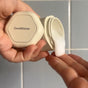 A person dispenses creamy white conditioner from a Cadence Hair Duo Travel Container onto their fingers against a tiled bathroom wall.