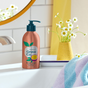 A bottle of Grove Co. Hand Soap sits on the bathroom sink near a yellow vase with white flowers. A colorful towel is nearby, and a round mirror reflects the charm of plant-based ingredients in its recyclable aluminum packaging against the white tiled wall.