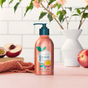 A copper Grove Co. Hand Soap bottle with a pump sits on pink, with a peach slice, coral cloth, and pink flowers in a white vase against white tiles, highlighting its plant-based formula and recyclable aluminum packaging.