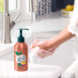 A person uses foamy hand soap at the kitchen sink. On the counter, a bottle of Grove Co. Hand Soap with a pump dispenser, crafted from plant-based ingredients, sits nearby. In the blurred background, charming kitchen elements and a fruit bowl enhance the scene.