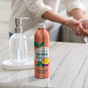 A bottle of Grove Co. Hydrating Hand Soap Refill, made from plant-based ingredients, sits on a marble countertop next to a clear glass soap dispenser, while someone washes their hands in the background.