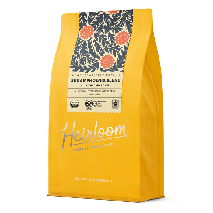 A yellow 12oz bag of Heirloom Coffee Roasters Sugar Phoenix ROC Whole Bean Coffee features a dark label with orange berries and leaves, offering a light-medium roast regenerative organic coffee with almond butter, berry, and floral notes.