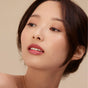 A woman with smooth, glowing skin and natural makeup wears Henne Organics Luxury Lip Tint for glossy, hydrated lips. Her soft brown eyeshadow and neat brows complement her look, while her dark hair is loosely pulled back as she gazes upward.