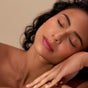 A woman with curly dark hair and glowing skin rests her head on her hand, eyes closed, wearing natural makeup and Henne Organics Luxury Lip Tint for a hint of hydrating color against a neutral background.