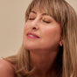 A middle-aged woman with light brown hair and bangs smiles gently with her eyes closed, wearing Henne Organics Luxury Lip Tint, looking relaxed and content against a soft beige background.