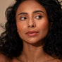 A woman with curly black hair and glowing skin looks to the side, wearing natural makeup, defined brows, and a subtle smile. She showcases Henne Organics Luxury Lip Tint against a neutral background.