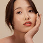 A woman with radiant, smooth skin rests her face on her hand, wearing Henne Organics Luxury Lip Tint—a hydrating lip color that gives her lips a soft, natural peach tone and completes her calm, thoughtful look.