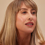 A woman with straight blonde hair and bangs looks slightly to the side, wearing light makeup and Henne Organics Luxury Lip Tint in pink. She is posed against a neutral background.