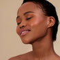 A woman with smooth, glowing skin and closed eyes smiles softly, wearing Henne Organics Luxury Lip Tint as she tilts her head back against a plain beige background.