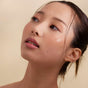 A woman with smooth, glowing skin looks upward, her softly blushed cheeks and hydrated lips enhanced by Henne Organics Luxury Lip Tint. Dark hair pulled back, wisps frame her face against a neutral background.