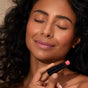 A woman with curly hair and radiant skin smiles softly with her eyes closed, holding a Henné Organics Luxury Lip Tint to her lips, highlighting the hydrating color from Henne Organics.