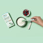 A hand pours Hilma Kids Happy Tummy Fiber Powder Packets into a bowl of yogurt with a spoon. Beside the bowl are berries and the Hilma box, all arranged on a pastel green background.