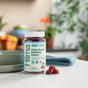 A bottle of Hilma Kids Happy Tummy Regularity Support Gummies rests on a kitchen counter with several gummies beside it, while blurred fruit and other kitchen items appear in the background.