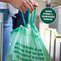 A person pulls the drawstring handles of a HoldOn Bags Compostable Tall Kitchen Bag (13 gal, Unscented) near a trash bin. A circular label reads SUPER STRONG 💪 DRAWSTRING HANDLES. These green BPA-free garbage bags feature printed text.