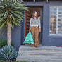 A woman on a front porch holds HoldOn Bags' Compostable Tall Kitchen Bags, 13 gal - Unscented in a green reusable tote. She wears a striped sweater and tan pants. Large plants and a scooter are near the steps of the modern, gray house behind her.