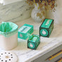 A marble countertop holds a white trash bin lined with Compostable Small Space Bags, two boxes of HoldOn Bags (one open to show a green bag roll), and dried flowers in a vase.