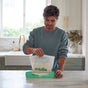 A man in a blue sweatshirt stands in a bright kitchen, pulling a HoldOn Bags Compostable Gallon Bag—freezer safe—from a green box on a white marble countertop.