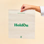 A hand holds up a translucent HoldOn Bags Compostable Gallon Bag—freezer safe, BPA free, and labeled in green—against a beige background. The bag features a zipseal, dotted line, and composting instructions near the top.