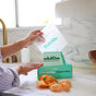 Someone puts peeled orange segments into a clear, compostable sandwich bag from HoldOn Bags on the kitchen counter, next to a green box labeled “A Better Way, A Better Bag.” These freezer-safe bags are food-safe and eco-friendly.
