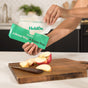 A person pulls a Compostable Snack Bag - Freezer Safe by HoldOn Bags from a green box labeled “A Better Way, A Better Bag” while apple slices and a knife sit on a cutting board on the kitchen counter.