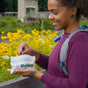 A woman outdoors, smiling by a field of yellow flowers, holds HoldOn Bags’ Compostable Snack Bags—freezer safe and eco-friendly—while wearing a purple shirt and gray backpack.