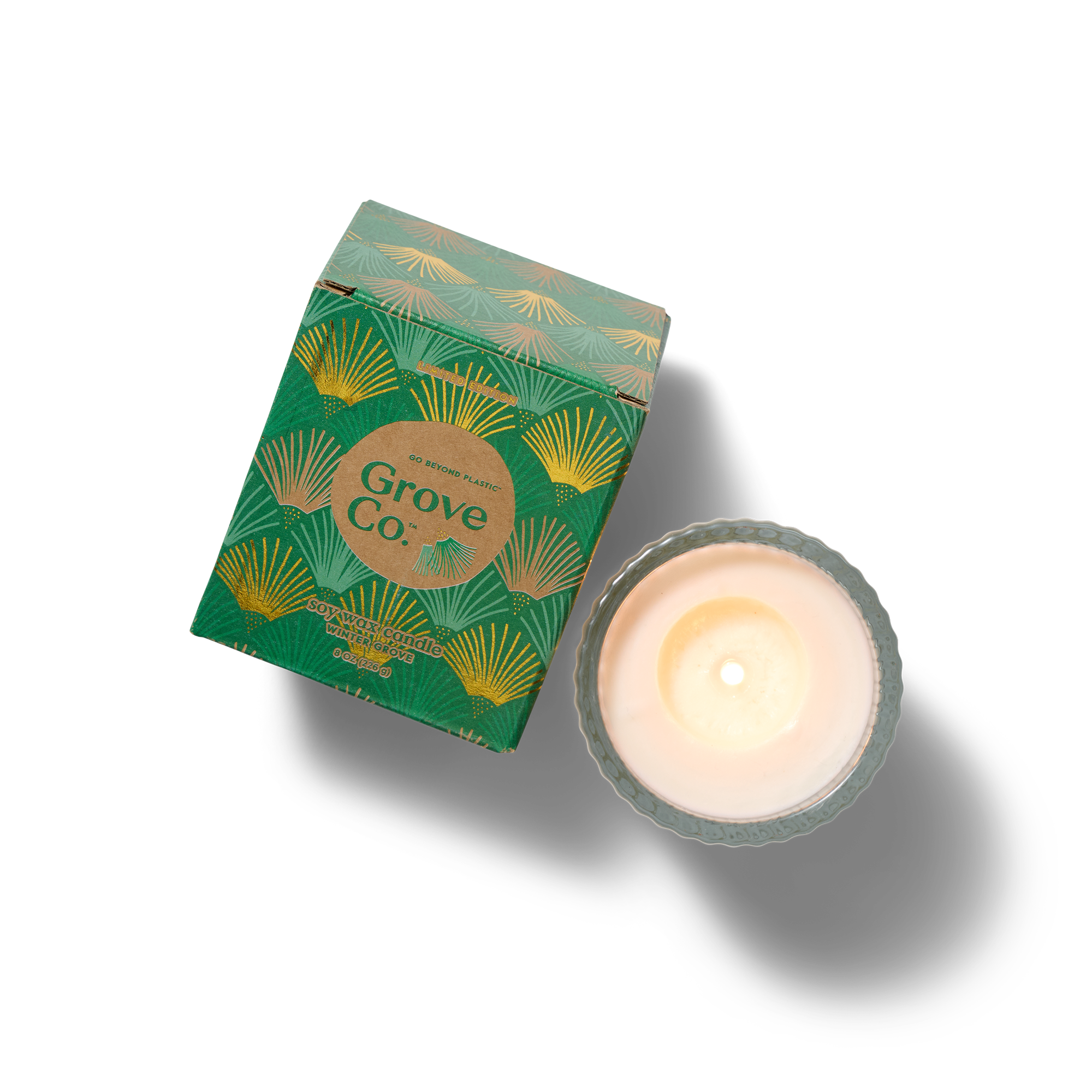 A green, patterned Grove Co. candle box is positioned next to a lit white candle in a decorative glass holder, both on a white background.