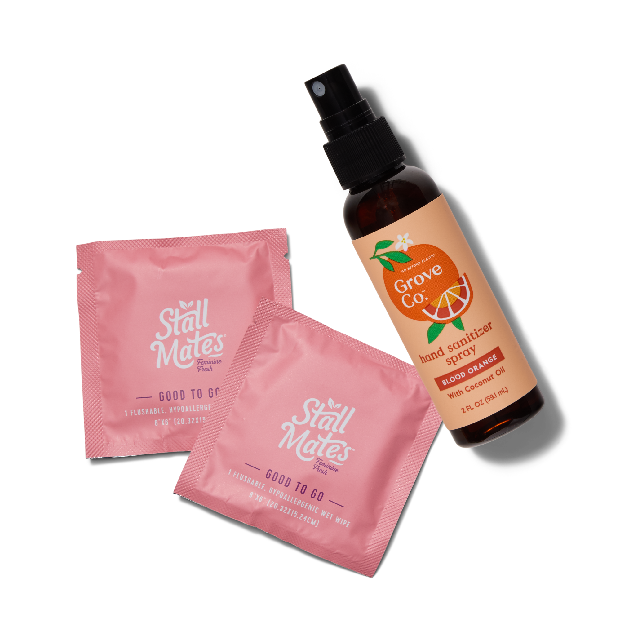 Two pink Stall Mates flushable wipes packets are next to a brown spray bottle of Grove Co. Blood Orange hand sanitizer with an orange and grapefruit illustration on the label, all on a white background.