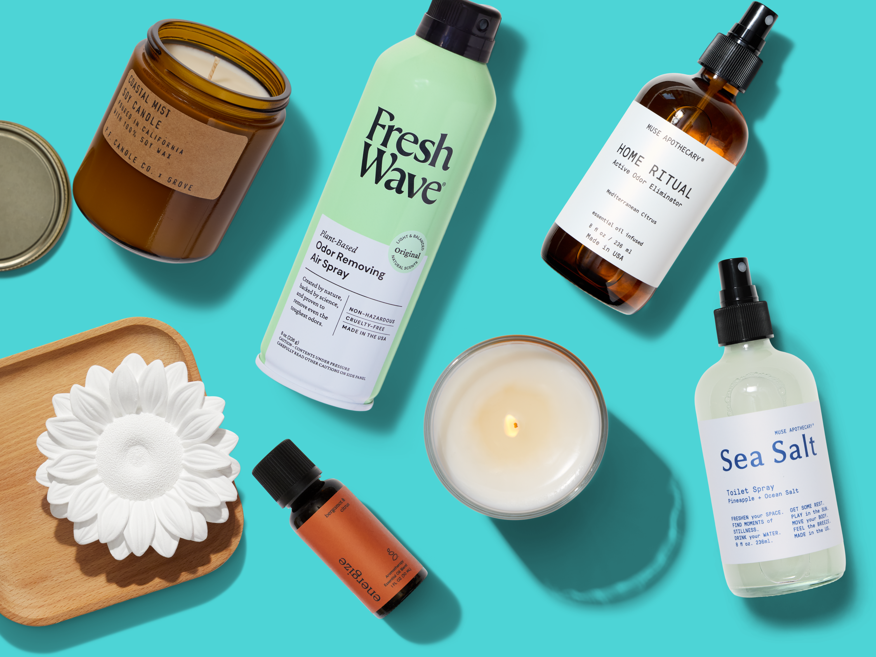 A flat lay image of various home fragrance products, including candles, spray bottles, a dropper bottle, a diffuser flower, a small vial, and a wooden tray on a bright turquoise background.