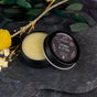 An open tin of Chagrin Valley Soap & Salve Honey Butter Lip Balm sits on a dark stone surface among eucalyptus leaves and dried yellow flowers. The organic balm appears pale and smooth inside the black container with a decorative lid.