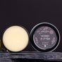 An open Chagrin Valley Soap & Salve Honey Butter Lip Balm tin rests on rustic wood, revealing its smooth balm. The round lid shows floral designs and reads, “Honey Butter Lip Balm with Real Honey”—ideal for soothing dry, chapped lips.