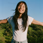 A person with long dark hair smiles joyfully outdoors, arms outstretched in a sunny green landscape—embracing confidence and comfort with Ursa Majors Hoppin Fresh Deodorant under a clear blue sky.