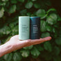 A hand holds two Ursa Major deodorant sticks—Hoppin’ Fresh (light green) and Forest Fix (dark green)—against a leafy backdrop, showcasing their essential oil blends.