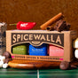 A box of Spicewalla Hot Chocolate Drink Mix Collection is surrounded by chocolate pieces, a red chili pepper, cinnamon sticks, and marshmallows on a green tile surface.